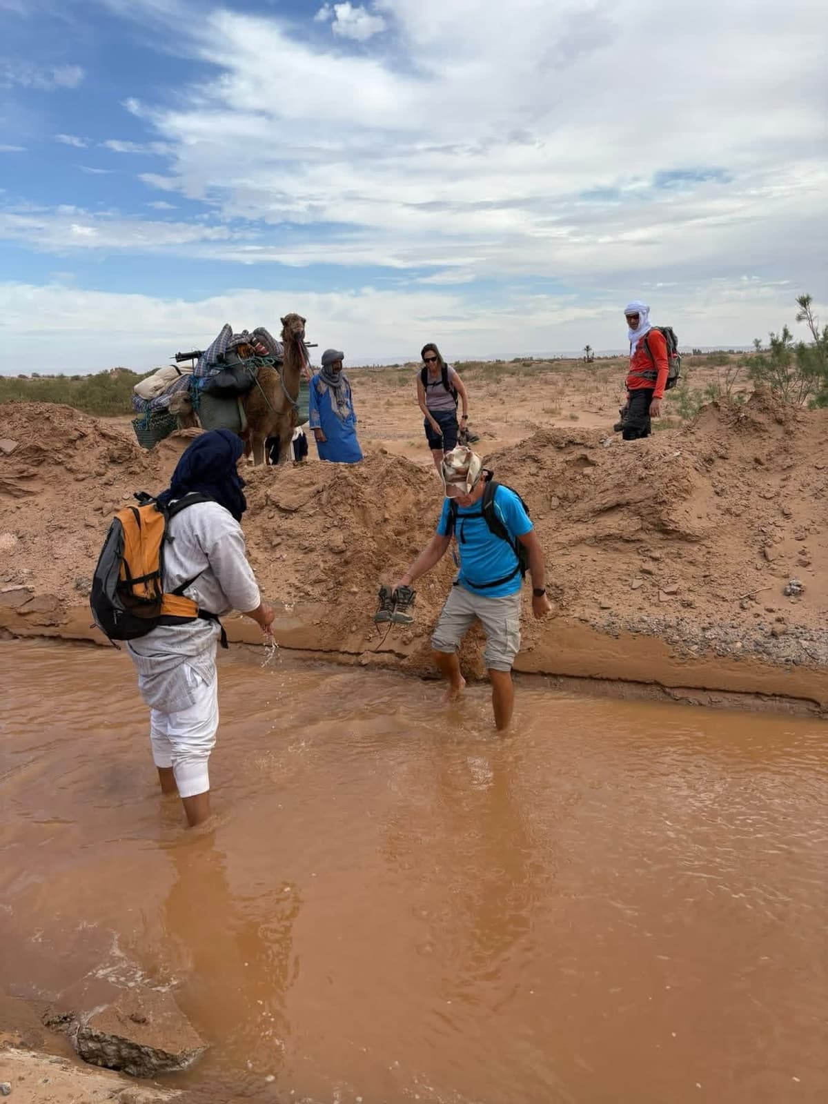 Travelers enjoying a Morocco walking tour through a desert landscape, crossing shallow water with local guides and camels