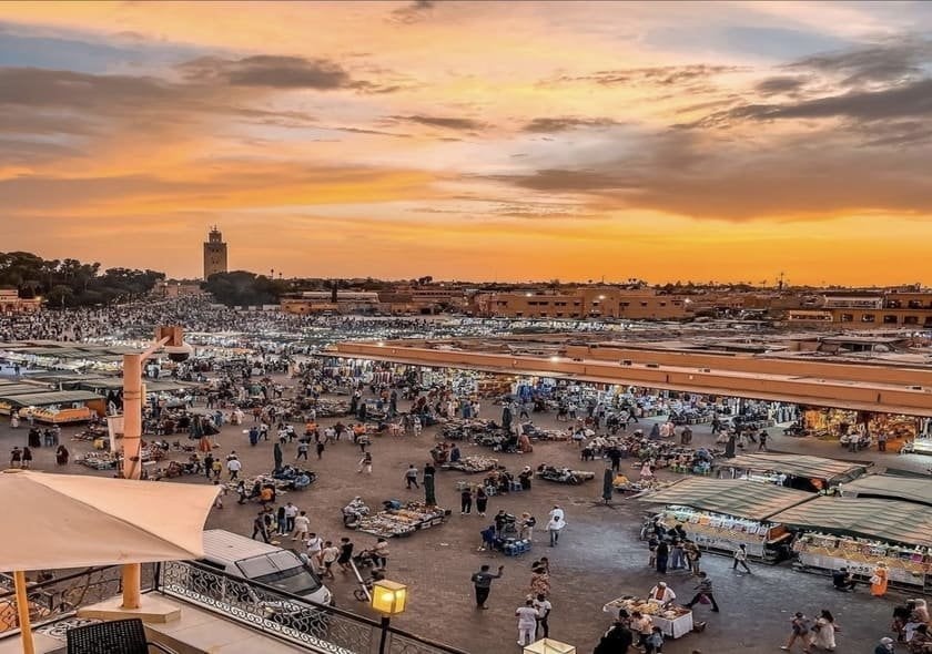 Panoramic view of Jemaa el-Fnaa Square in Marrakech, Morocco, showing bustling market stalls, crowds of people, rooftop cafés, and a golden sunset over the Medina